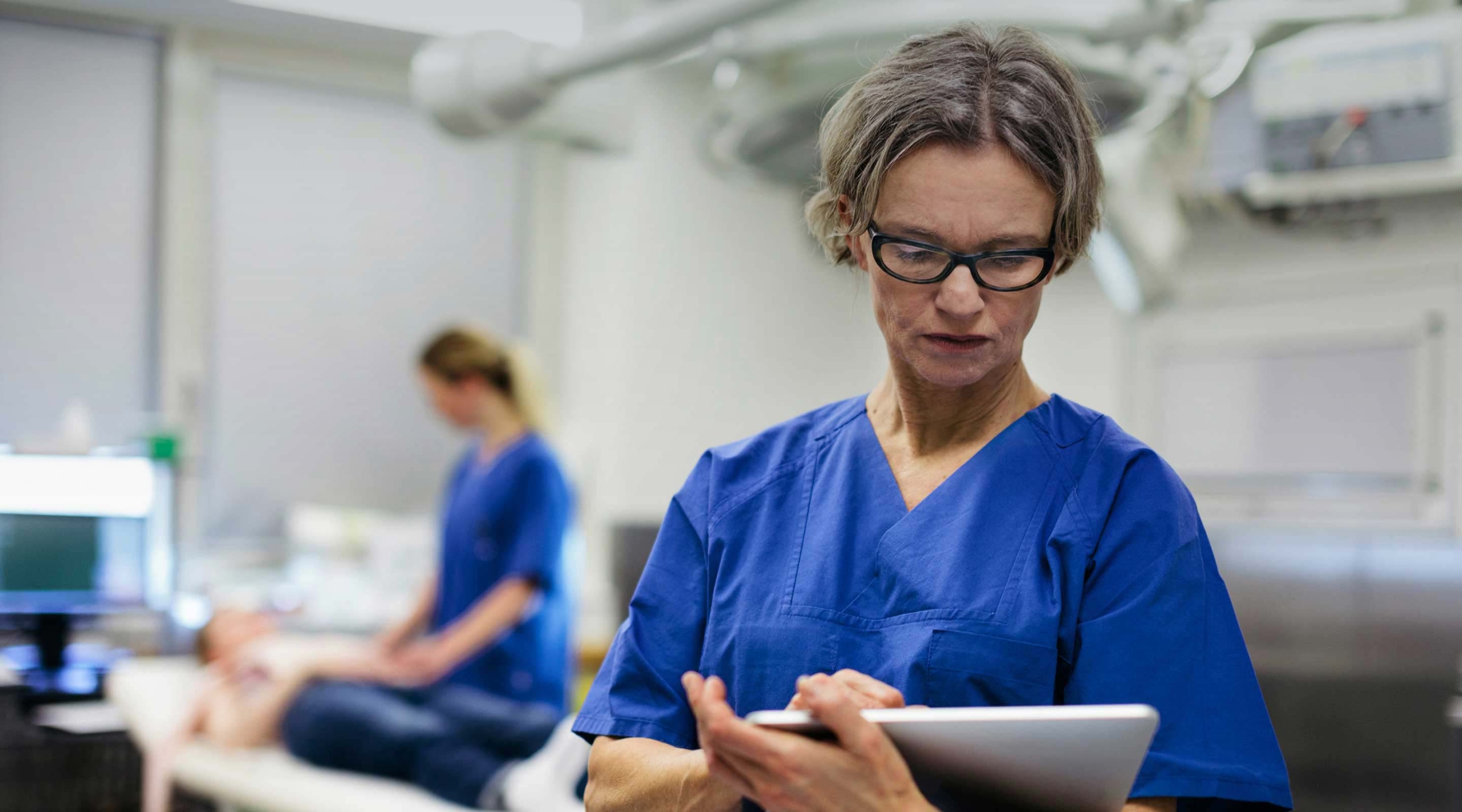 Healthcare professional in blue scrubs using a tablet while standing in a modern clinical treatment room, with a colleague caring for a patient on an examination bed in the background.